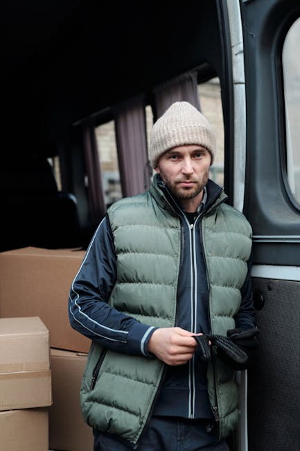 A man and a woman are engaged in a home relocation process involving furniture transport and packing, standing beside an open moving van parked on a paved driveway. The man, dressed in a white polo sh