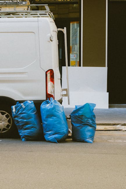 A white commercial van is parked on the pavement outside a building, with its rear doors closed and part of the side visible, including a vertical red tail light. Three large blue plastic bags filled with bulky waste materials are positioned on the ground next to the van, likely prepared for disposal or removal during a home relocation process. The bags appear to contain household items or debris, possibly from packing or clearing out a property. Behind the van, the building features a glass window and doorway, partially framed with metal, indicating an entrance area where furniture or packing supplies might be moved in or out. The scene suggests an ongoing loading or disposal operation as part of packing and moving activities, with the environment illuminated by natural light. The presence of this waste collection aligns with services provided by companies like Man and Van Paddington, focused on removals, packing, and efficient logistics for house relocations or decluttering tasks.