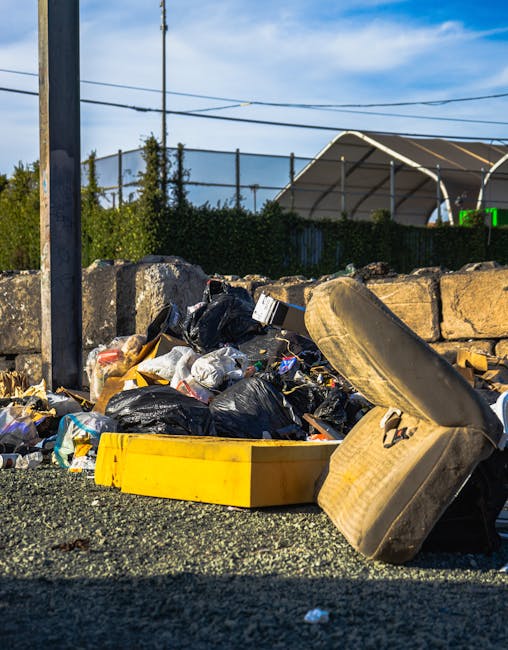 A pile of mixed waste materials, including black plastic trash bags, a yellow plastic storage container, a large old fabric-covered armchair, and various pieces of rubbish, are situated outdoors on a gravel surface next to a stone wall. In the background, there are green bushes, a metal fence, and a small building with a sloped roof under a partly cloudy sky. The scene appears to be an area designated for disposing of bulky waste, relevant to home relocation or clearance services. Occasionally, Man and Van Paddington's removals team might collect such items during furniture transport or packing and moving processes, as part of their house removal operations.