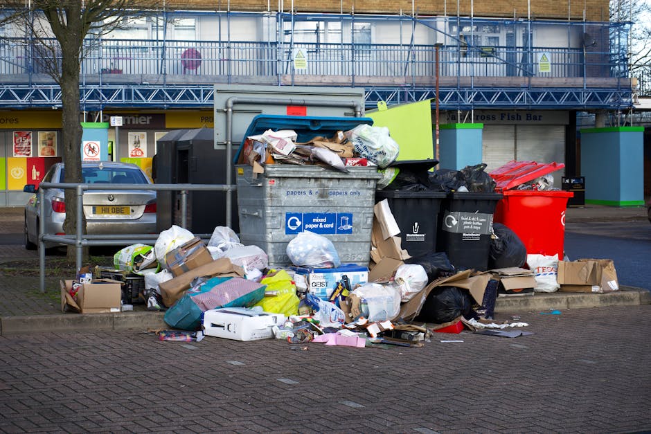 A white commercial van is parked on the pavement outside a building, with its rear doors closed and part of the side visible, including a vertical red tail light. Three large blue plastic bags filled with bulky waste materials are positioned on the ground next to the van, likely prepared for disposal or removal during a home relocation process. The bags appear to contain household items or debris, possibly from packing or clearing out a property. Behind the van, the building features a glass window and doorway, partially framed with metal, indicating an entrance area where furniture or packing supplies might be moved in or out. The scene suggests an ongoing loading or disposal operation as part of packing and moving activities, with the environment illuminated by natural light. The presence of this waste collection aligns with services provided by companies like Man and Van Paddington, focused on removals, packing, and efficient logistics for house relocations or decluttering tasks.
