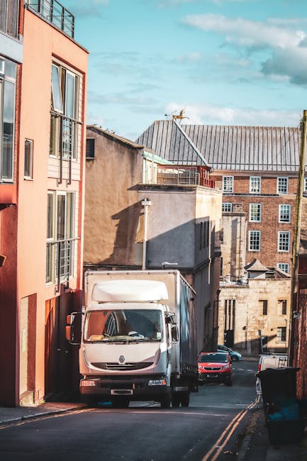 A white delivery van is parked on a narrow urban street, positioned closely next to a multistory pink and beige residential building on the left side of the image. The van's front windshield and side mirrors are visible, with the vehicle partially blocking a small section of the street. Behind the van, there are additional parked cars, including a red vehicle further down the road. The street is lined with older buildings constructed from brick and concrete, featuring small windows and flat roofs, with some structures showing signs of weathering. The road surface is asphalt with double yellow lines along the curb, and a black wheelie bin is placed on the sidewalk near the corner. Shadows cast by the buildings suggest late morning or early afternoon lighting. This scene illustrates a typical location where house removals or furniture transport might take place, with commercial vehicles like those operated by Man and Van Paddington prepared for home relocation and moving services.