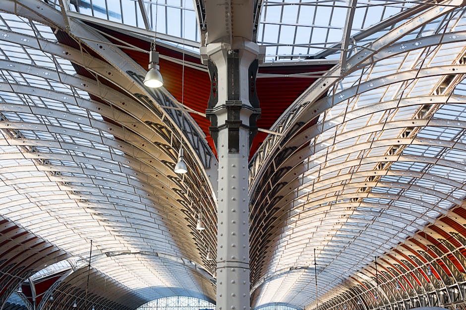 A detailed view of the interior ceiling structure of a large, modern transportation hub or warehouse with an arched, ribbed metal roof supported by central steel beams. The ceiling is composed of overlapping, semi-transparent panels that allow natural light to filter through. Several hanging industrial-style lights are suspended from the ceiling beams, providing additional illumination. The spacious environment features a high, curved roof design often associated with large-scale logistics facilities used for home relocation or furniture transport. The image captures the intricate metal framework and the expansive, open interior characteristic of buildings used to facilitate moving and packing services, such as those provided by Man and Van Paddington.