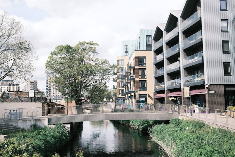 A view of a modern urban residential area adjacent to a small river, with a pedestrian bridge crossing over the water. The scene shows a collection of contemporary multi-storey apartment buildings with balconies, some constructed with grey and beige cladding materials. On the pavement, there is a stack of cardboard boxes and plastic-wrapped furniture, positioned near the building entrance, indicating a packing or home relocation process. Nearby, a person is seen lifting a cardboard box onto a trolley for transport, inside or just outside the property doorway. The environment is well-lit with natural daylight, emphasizing the street scene and the nearby greenery, including a large leafy tree and bushes along the riverbanks. The adjacent buildings and the river landscape reflect typical elements involved in furniture transport and packing logistics, relevant to the house removals services offered by Man and Van Paddington, as seen on the webpage for Paddington Basin student moves, flats, lifts, and access.