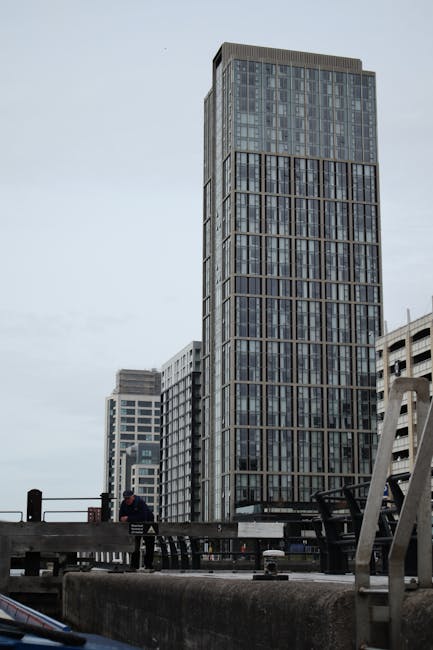 A tall modern high-rise building with a glass facade and grid-like structure, standing amidst other commercial buildings in an urban area under an overcast sky. The foreground shows part of a flat rooftop with a low parapet, black railings, and some equipment or structures. A person wearing dark clothing is seen near the edge of the rooftop, possibly involved in a home relocation or moving process. The scene indicates an urban environment suitable for house removals and furniture transport, with the building's height and surrounding architecture highlighting the cityscape context, consistent with the services provided by Man and Van Paddington for flats, lifts, and access during moving and packing activities.