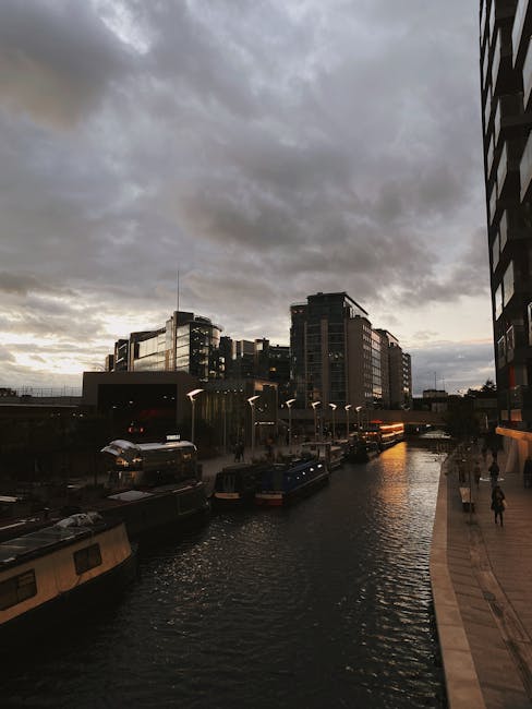 A view of a modern urban residential area adjacent to a small river, with a pedestrian bridge crossing over the water. The scene shows a collection of contemporary multi-storey apartment buildings with balconies, some constructed with grey and beige cladding materials. On the pavement, there is a stack of cardboard boxes and plastic-wrapped furniture, positioned near the building entrance, indicating a packing or home relocation process. Nearby, a person is seen lifting a cardboard box onto a trolley for transport, inside or just outside the property doorway. The environment is well-lit with natural daylight, emphasizing the street scene and the nearby greenery, including a large leafy tree and bushes along the riverbanks. The adjacent buildings and the river landscape reflect typical elements involved in furniture transport and packing logistics, relevant to the house removals services offered by Man and Van Paddington, as seen on the webpage for Paddington Basin student moves, flats, lifts, and access.