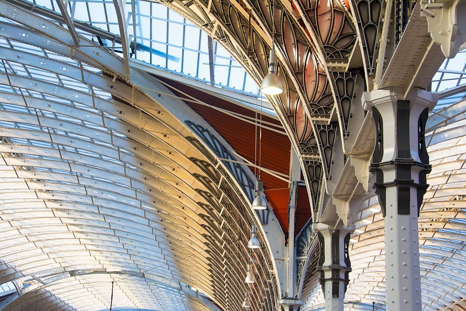 An interior view of Paddington Station's ornate metal roof structure, featuring curved metal beams painted in white and black, supporting a semi-circular glass skylight that allows natural light to illuminate the space. Several hanging pendant lights are suspended from the ceiling, enhancing the lighting of the station's high, arched roof. The detailed metalwork includes riveted joints and intricate decorative elements, reflecting Victorian-era architectural design. While the image does not show furniture or moving equipment, it depicts the station environment where home relocation and furniture transport operations may take place, often undertaken by companies like Man and Van Paddington, especially during packing and loading processes for house removals.