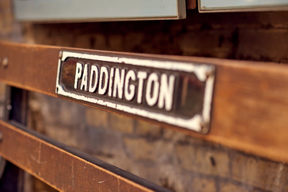 Close-up image of a vintage metal and wood sign with the word 'PADDINGTON' in bold, white uppercase letters on a black background, mounted on a wooden panel. The sign shows signs of wear and rust, indicating age or outdoor exposure. The wooden panel behind the sign is dark brown with visible grain and texture. The photo is taken at an angle, highlighting the weathered appearance of the sign and wood. This image reflects the context of house removals or transport logistics near Paddington Station, captured by Man and Van Paddington as part of their removals services, emphasizing the need for careful handling and parking considerations when relocating belongings or furniture in the area.