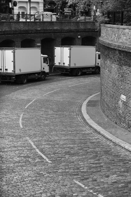 A black-and-white photograph showing two box trucks parked on a curved cobblestone street beneath a bridge. The trucks, with closed cargo compartments, are positioned side by side facing away from the camera, while the street curves to the right and is marked with dashed white lines. To the right, there is a tall brick wall with a small street sign attached, and above the vehicles, a railing borders a higher-level area with trees and pathways. The scene appears to be in an urban environment suitable for home relocation and furniture transport, illustrating the loading process for removals by a professional service such as Man and Van Paddington, which facilitates efficient and secure packing and moving operations.