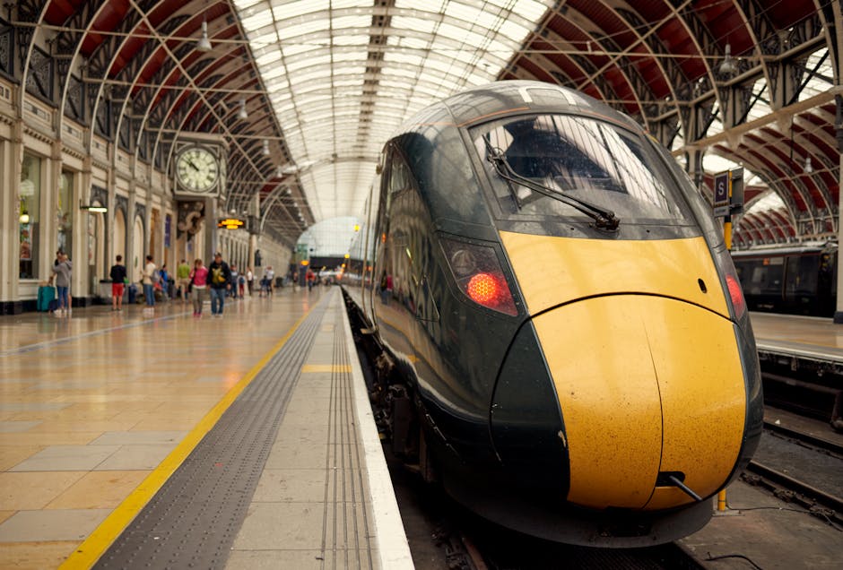 A modern high-speed train with a streamlined black and yellow exterior is positioned at a platform inside a historic train station with a curved, glass-paneled ceiling. The train's front nose is rounded, with windshield wipers and red indicator lights illuminated. The platform has a tactile strip along the edge and is made of light-colored tiles, with a yellow line marking the safety boundary. Several passengers are seen in the background, some standing and others sitting on benches, waiting near a large clock mounted on an ornate wall with classic architectural details. The station is well-lit, with natural light streaming through the glass ceiling, and features a spacious, airy environment suitable for home relocation logistics, including loading and unloading of luggage or furniture. The setting illustrates a busy transportation hub where moving and transport services, such as those provided by Man and Van Paddington, might coordinate parking permits and address moving logistics involving train transport within London.