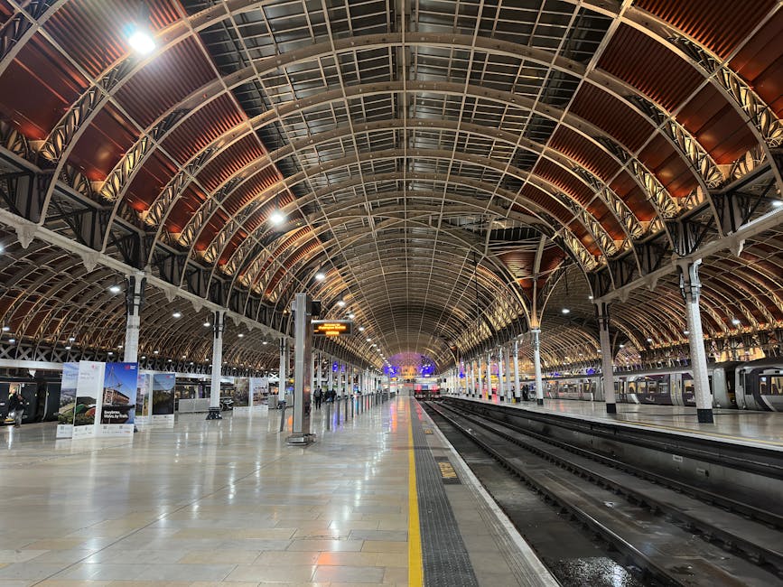 An expansive indoor train station with a high, arched ceiling constructed of metal and glass panels, illuminated by ceiling lights. The platform side features a row of classic black and white column-supported structures with bright signage and electronic departure boards overhead. Several travelers are visible in the distance, some walking along the platform with luggage, while others are waiting near the edge for arriving trains. On the right side, multiple high-speed train carriages with closed doors are stationary, ready for passenger boarding. The polished tiled floor reflects the lighting, and on the platform, there is a clear yellow safety line and tactile paving for accessibility. This setting illustrates the interior of a modern, well-lit railway station, suitable for handling home relocation or furniture transport logistics, especially as part of comprehensive house removals or packing and moving services provided by Man and Van Paddington.