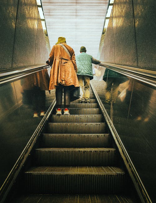Two individuals are riding an escalator inside a modern building, heading upward towards the light-filled ceiling. The person on the left is wearing a long, tan coat, dark pants, and red shoes, and is carrying a small bag. The person on the right is dressed in a green jacket and beige trousers, standing beside the first individual. The escalator's metal handrails and black steps are visible, with a reflective wall on each side. The environment indicates an indoor location with clean, smooth surfaces, and the bright lighting highlights the structured ceiling overhead. This scene exemplifies a typical home relocation setting, where moving materials and furniture might be transported through multi-story buildings, supporting the context of stair-only removals in W2 properties, as managed by Man and Van Paddington.