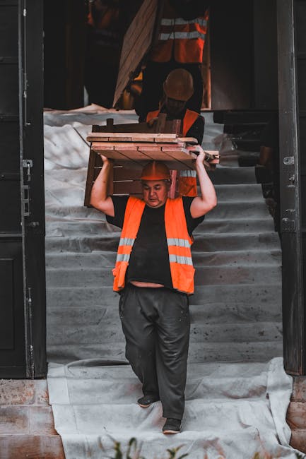 A man wearing an orange high-visibility vest and a hard hat is walking down a set of concrete stairs outside a building, carrying a bundle of wooden planks balanced on his shoulders. Behind him, another worker also dressed in safety gear is visible, descending further down the stairs with what appears to be additional packing or furniture items. The stairs are partially covered with a protective fabric sheet to safeguard against damage during the moving process. To the side of the stairs, there is a doorway leading into a property where furniture packing and relocation activities are taking place. The scene is illuminated by natural light, highlighting the careful handling of household items involved in a house removal or furniture transport operation, as part of a professional moving service by Man and Van Paddington, specialists in removals and home relocations, specifically addressing stair-only moves in W2 properties.